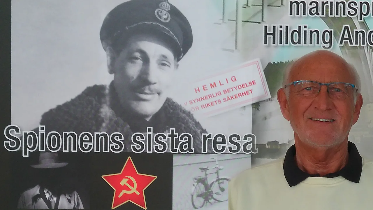 Göran Janrell framför en affisch för föreläsningen Spionens sista resa, med ett porträtt av Hilding Andersson och en HEMLIG-stämpel